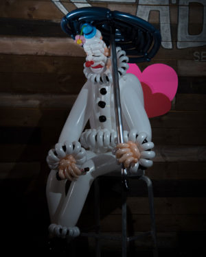 Sad Balloon clown in front of pink hearts holding an umbrella and empty beer can