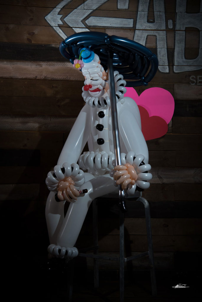 Sad Balloon clown in front of pink hearts holding an umbrella and empty beer can