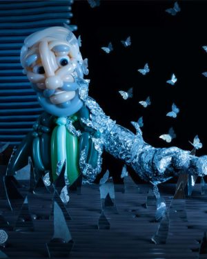 a shattered visage balloon sculpture of man with gray beard and green suit impaling his hand on broken mirror shard as tin foil envelops his arm with metal butterflies in background