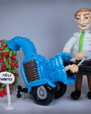 Balloon sculpture of boss in tie shoving worker into woodchipper wich is spitting out blood and dollar bills behind a help wanted sign