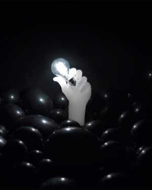 Reaching baloon sculpture wax hand reaching through sea of black balloon holding lit light bulb