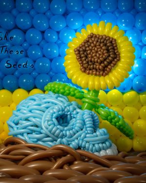 Take These Seeds balloon sculpture of a skull on the ground with a sunflower growing out of eye socket in front of ukranian flag