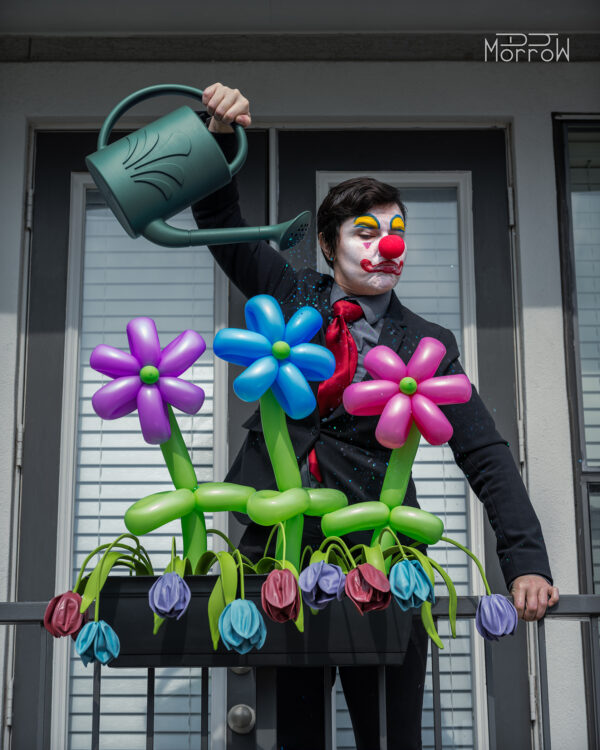 clown in business suit watering balloon flowers while more delicate flowers wither.