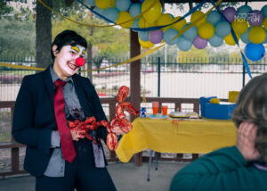 clown in business suit holding disemboweled intestines twisted into a balloon dog, grimacing and handing to a shocked child at birthday party