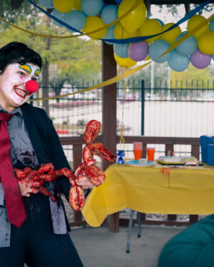 clown in business suit holding disemboweled intestines twisted into a balloon dog, grimacing and handing to a shocked child at birthday party