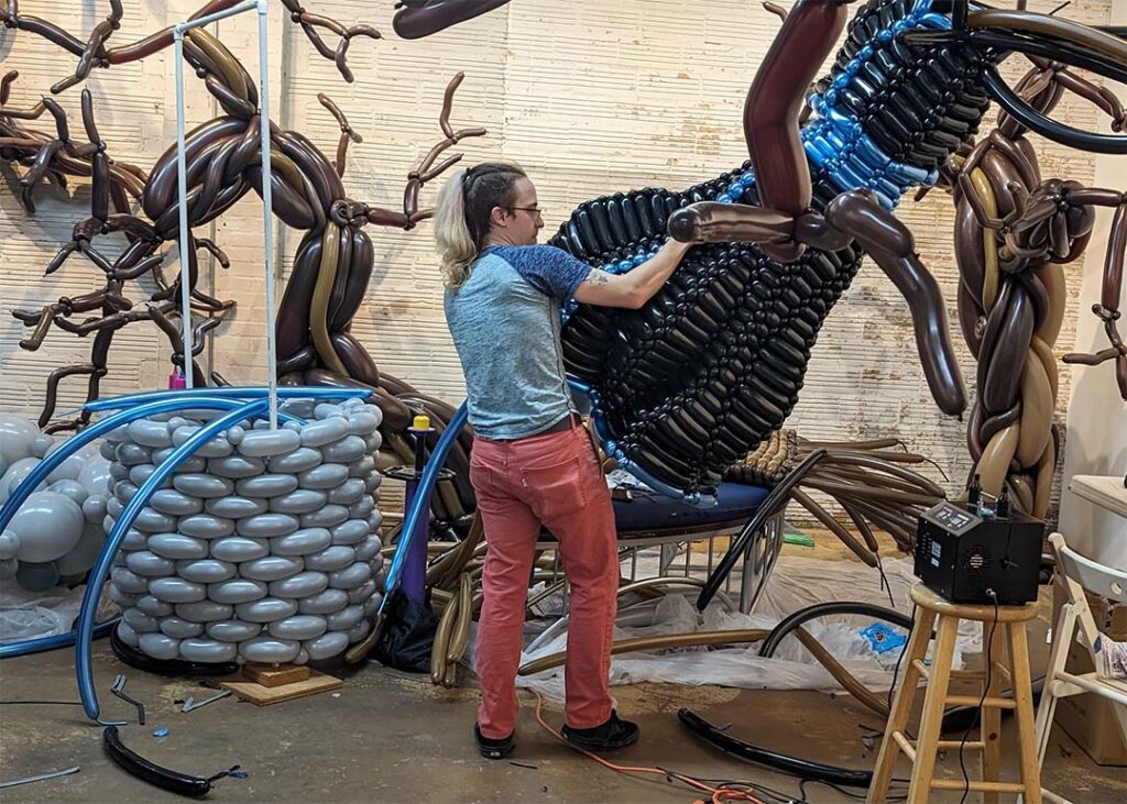 dj morrow artist building lightbringer balloon installation holding robes with tree in foreground and well in background